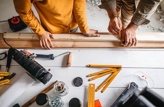 Two people measure wood for home improvements on a tool-covered table